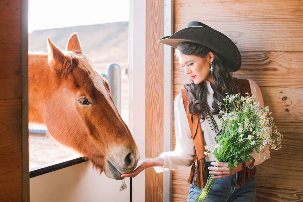 From 6th Collective x Dove Creek Ranch styled shoot image features a moden in leather vest, turquoise and silver jewelry, a bouquet of wildflowers, and a brown cowgirl hat feeding a horse through a stable door.